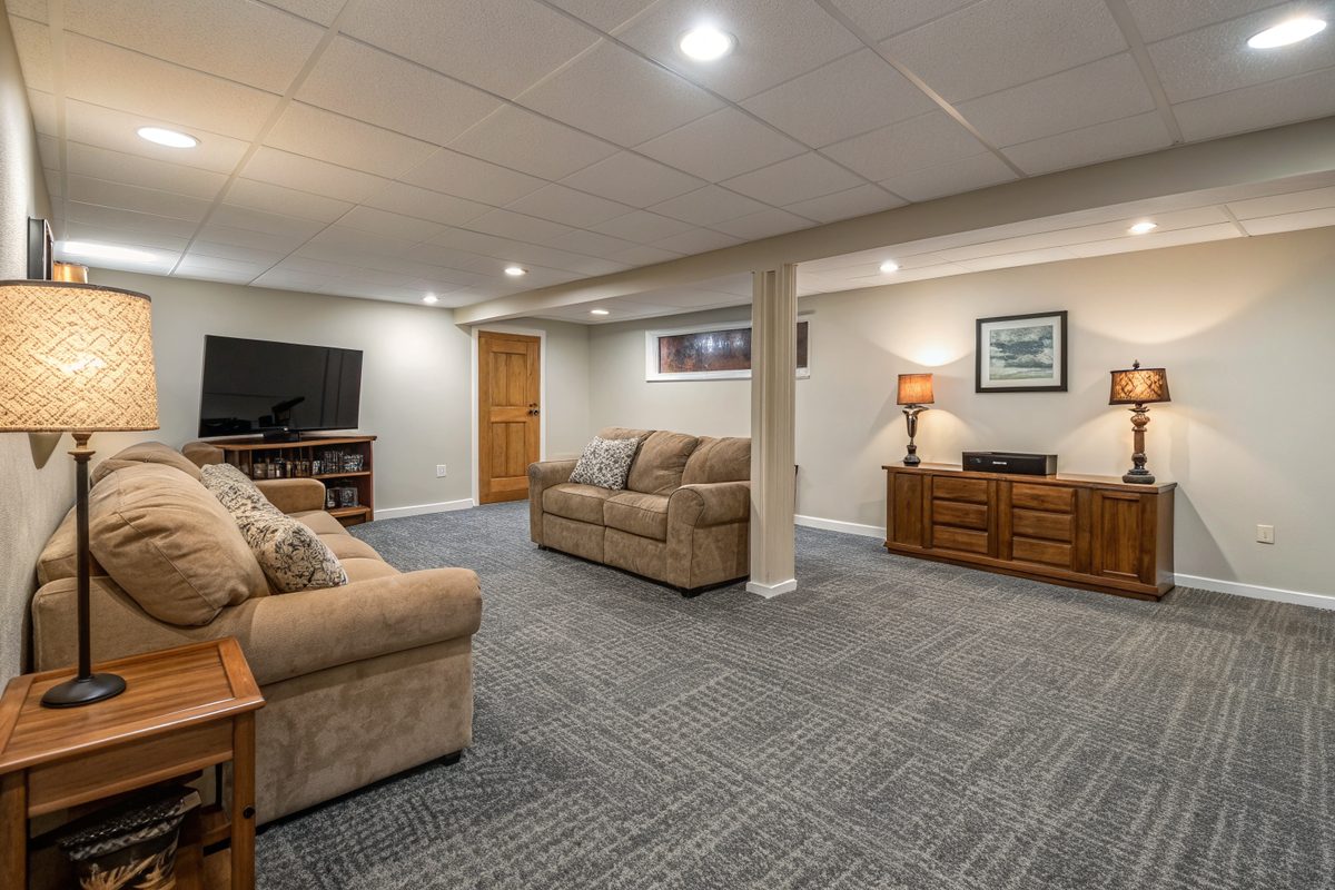 A dry finished basement family room with carpet tiles, a tan sectional sofa and warm lamps