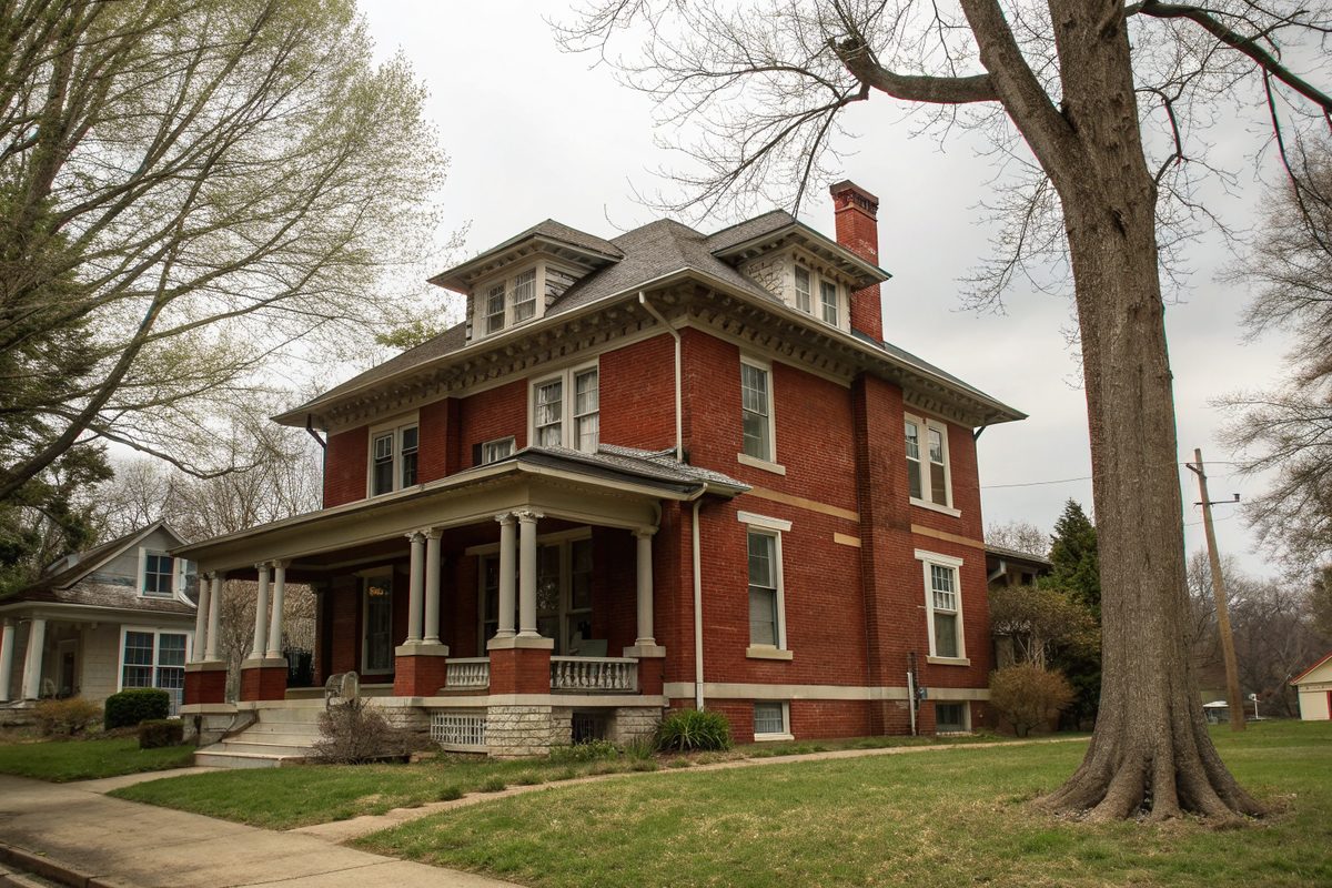 A two-story red brick foursquare house with a deep front porch and small basement windows on a limestone foundation